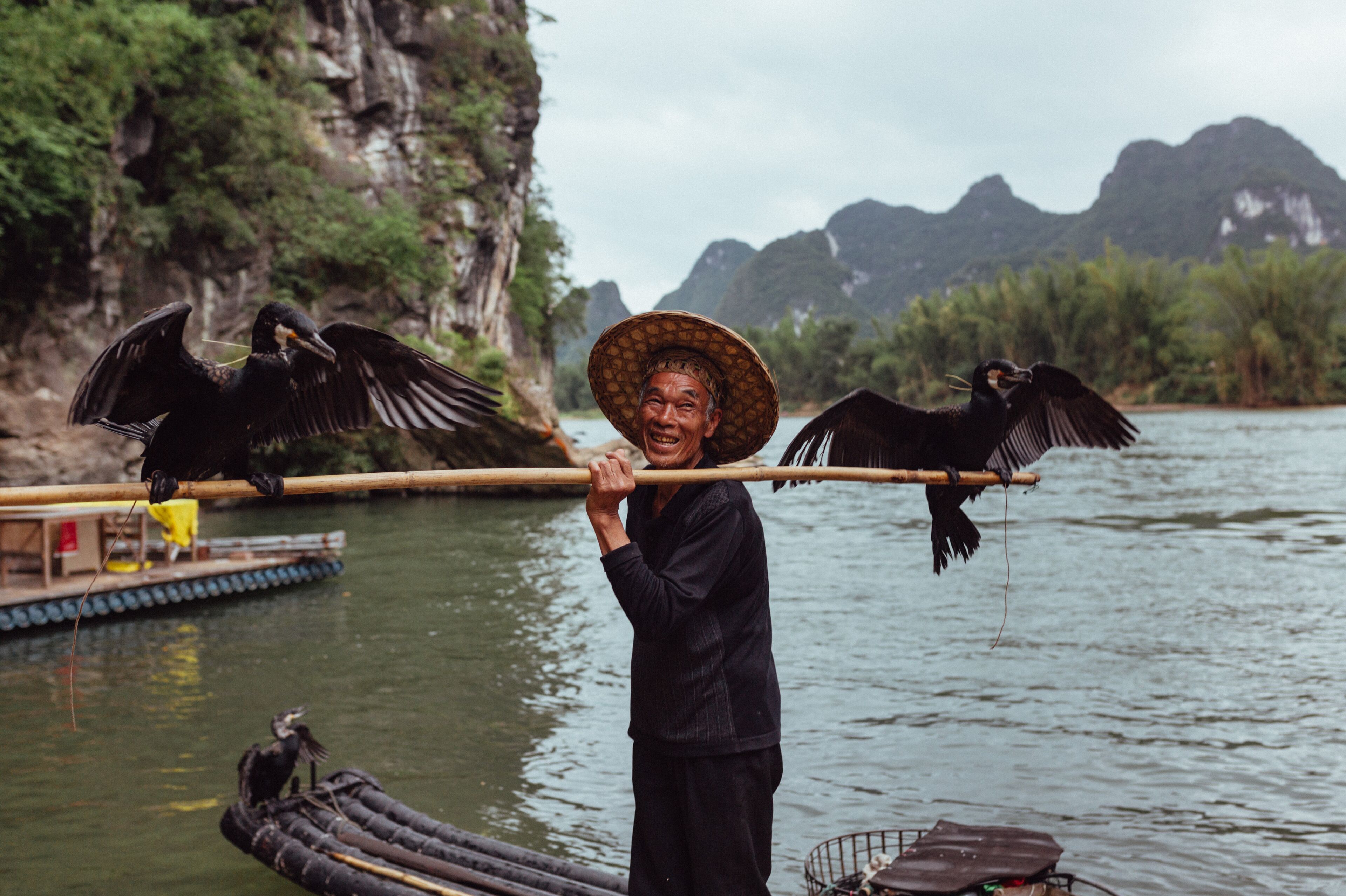 A person holding a long pole with two birds perched on it, standing by a body of water with boats and cliffs in the background.