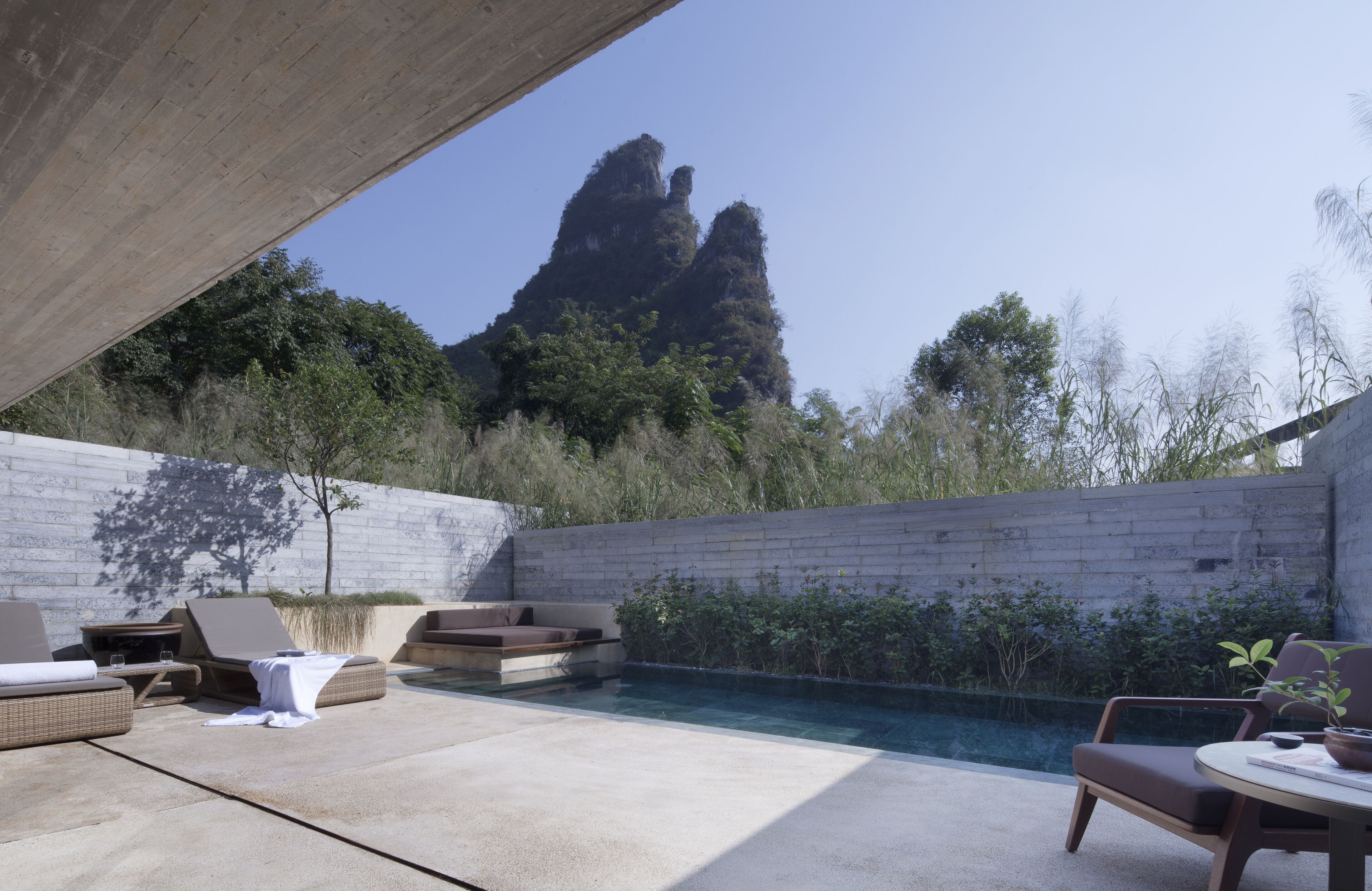 A pool area with lounge chairs and a table, surrounded by a concrete wall and vegetation.