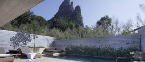 A pool area with lounge chairs and a table, surrounded by a concrete wall and vegetation.
