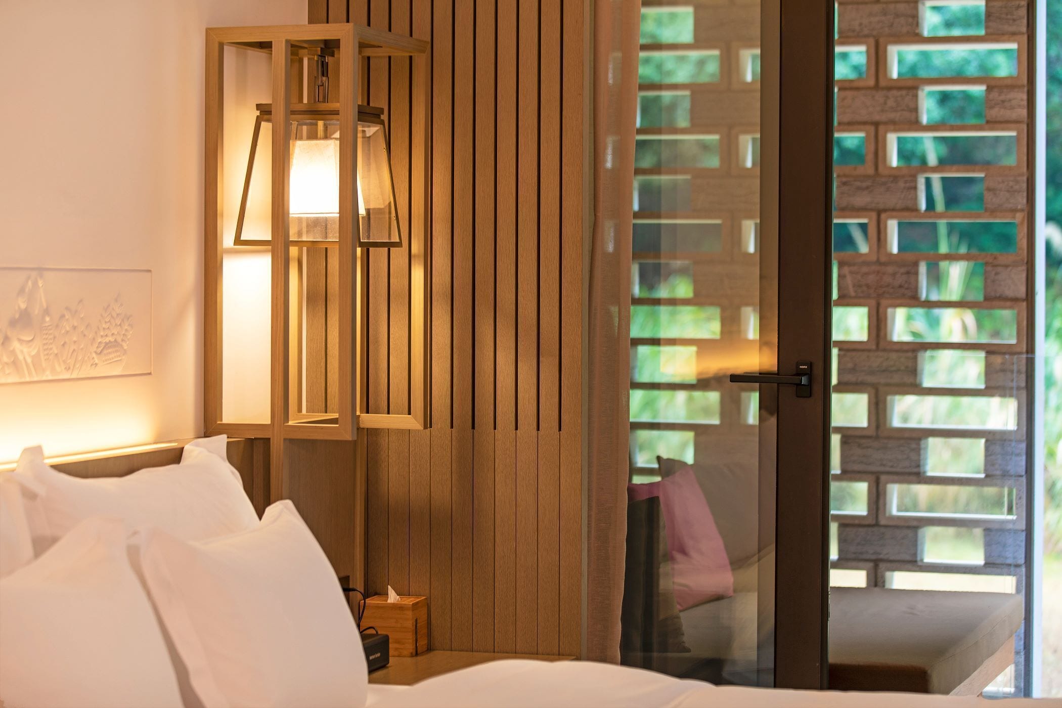 Habitación de hotel con cama, mesita de noche, lámpara y vistas a un espacio exterior con pared de ladrillo.