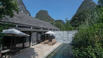A modern outdoor pool area with lounge chairs and umbrellas, surrounded by lush greenery and mountains in the background.