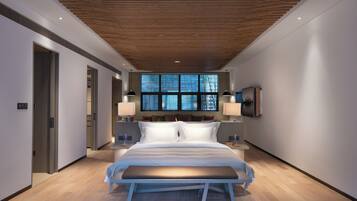A modern hotel room with a wooden ceiling, a large bed with white linens, a bench at the foot of the bed, and a large window with a view of trees.