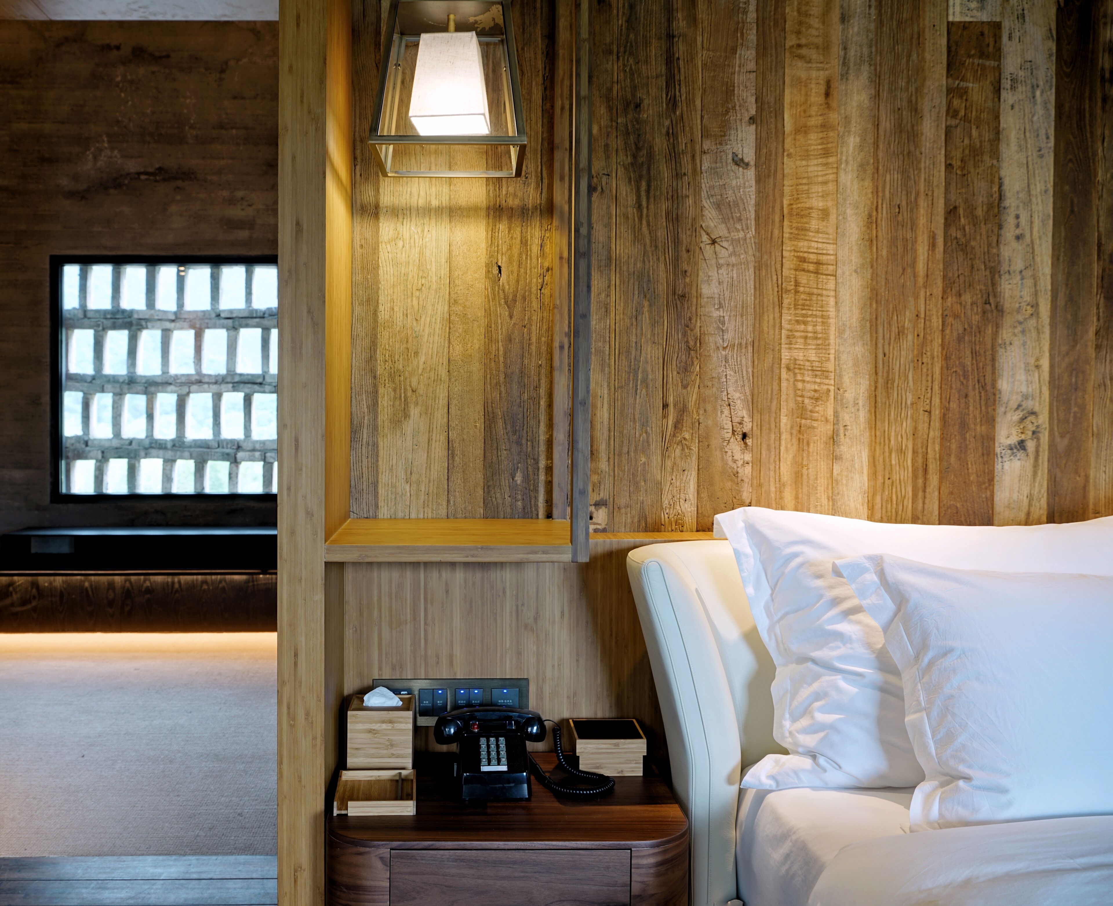 A hotel room with a wooden wall, a bed with white bedding, a bedside table with a telephone and a lamp, and a window with a view of a brick wall.