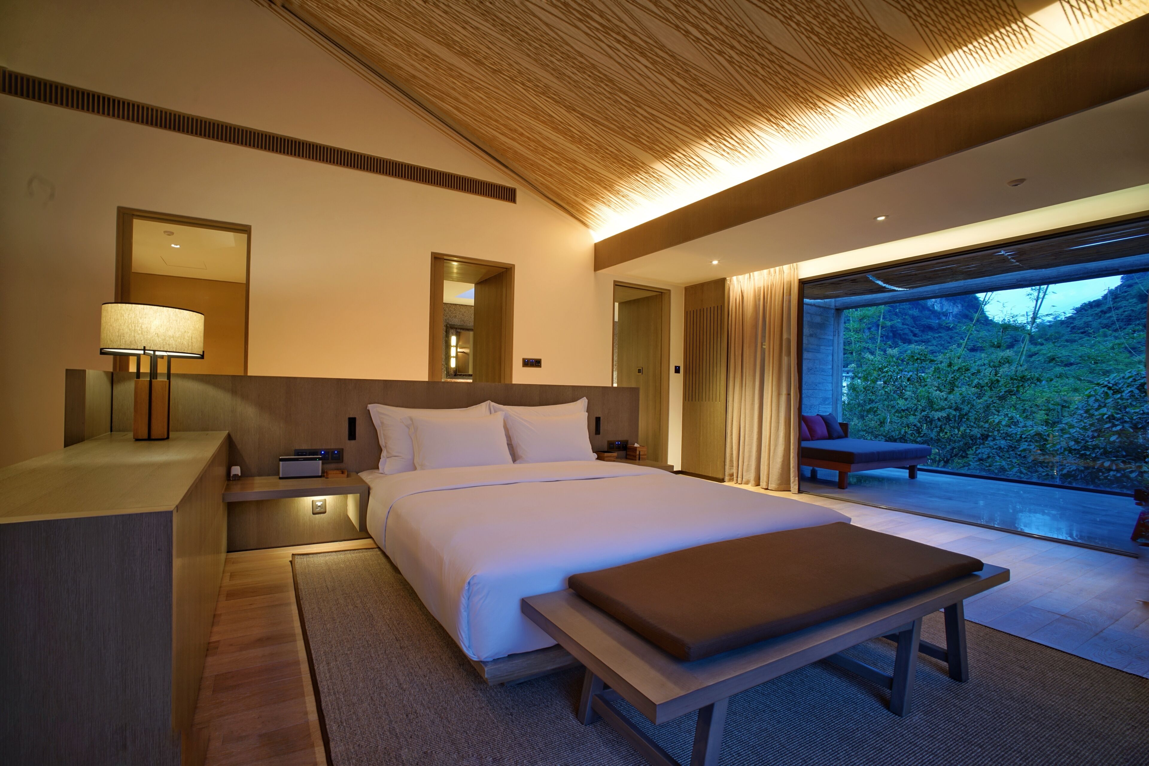 Habitación de hotel moderna con una cama grande, mesitas de noche, un banco y vistas al exterior.
