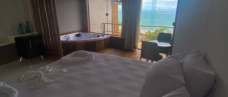 A bedroom with a large bed, a small desk, a chair, a bathtub, and a balcony with a view of the ocean.