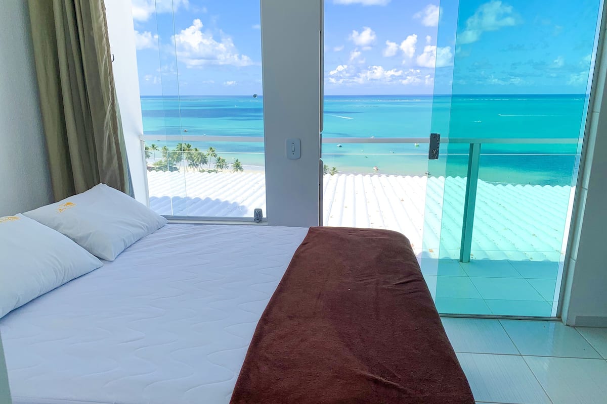 A bedroom with a bed, a brown blanket, and a view of the ocean through a glass door.
