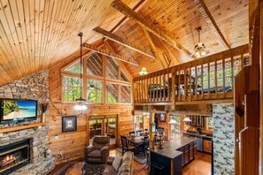 A spacious cabin with a stone fireplace, a large TV, a cozy seating area, and a kitchen with a high ceiling and wooden beams. - Smoky Mountain Lodge (Sevierville)