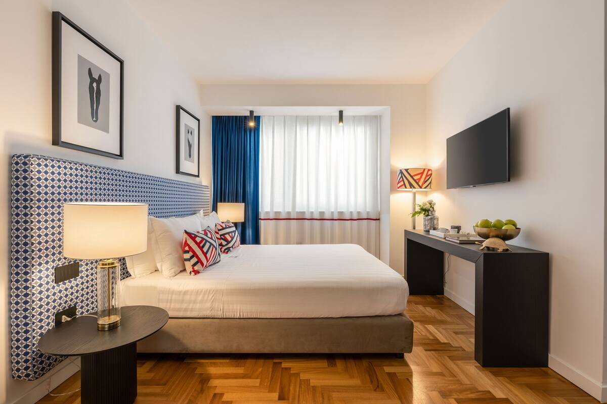 Una habitación de hotel moderna con un cabecero estampado, una cama con ropa blanca, una mesita de noche con lámpara, un televisor de pantalla plana y una pequeña mesita con adornos.