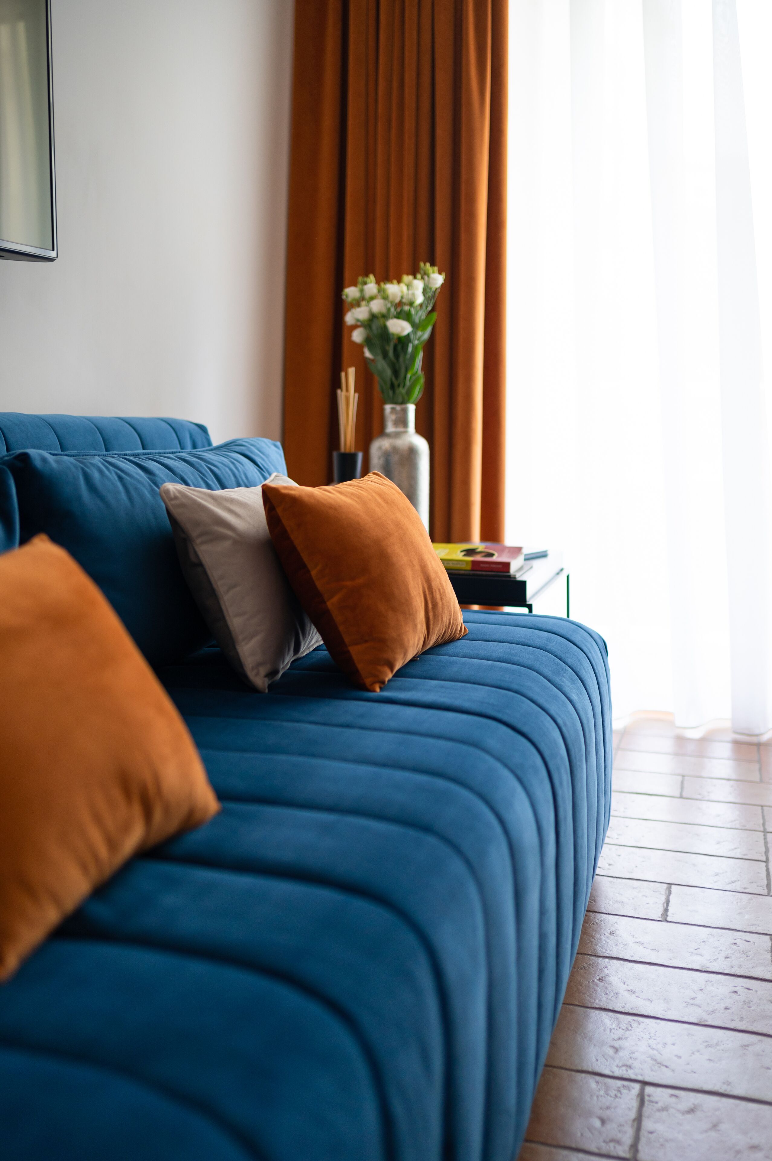 A blue sofa with multiple cushions, a small table with books and a vase of flowers, and a window with curtains.