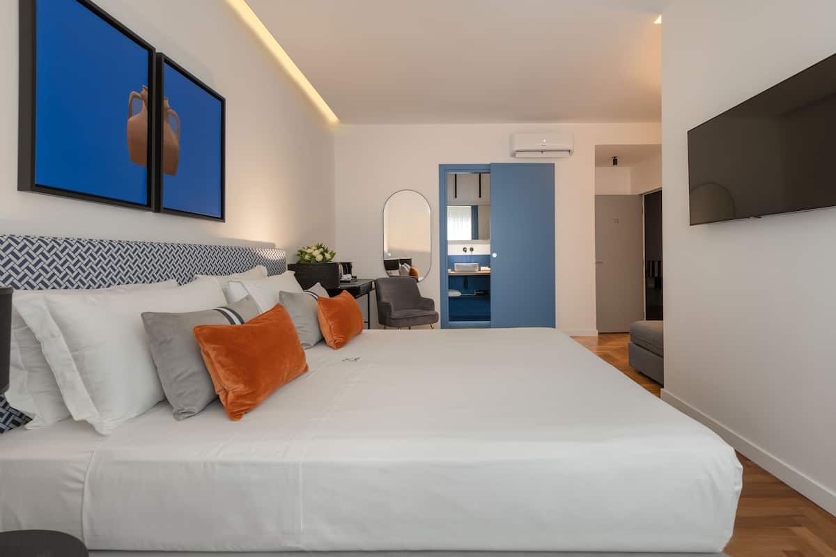 Habitación de hotel moderna con una cama grande, un escritorio, una silla, un televisor y un baño visible a través de una puerta abierta.