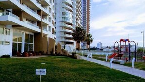A multi-story residential building with balconies, a grassy area with a playground, and a sign indicating a rental service. - Depto Costa Mansa (Coquimbo)