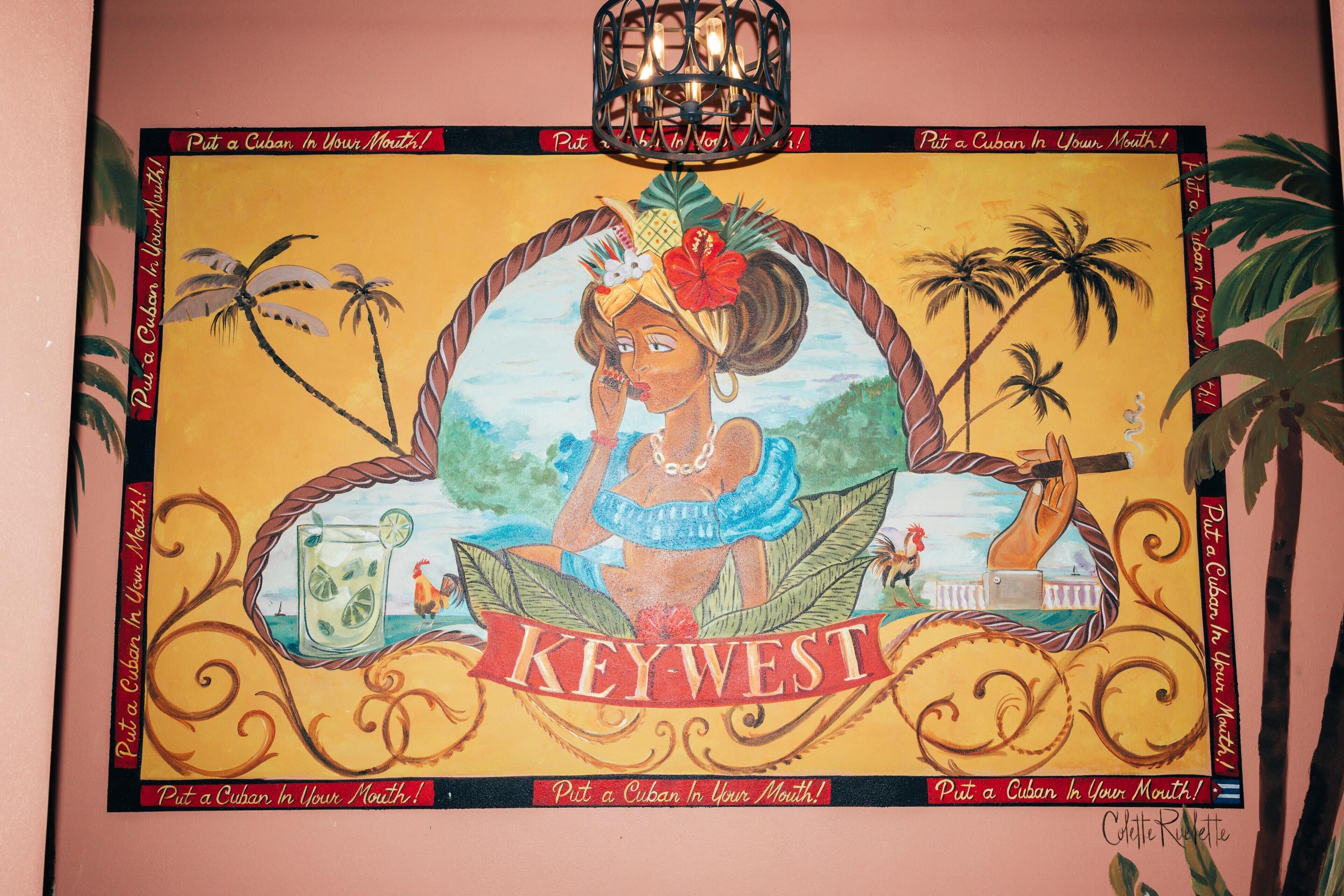 Une peinture aux couleurs vives, sur le thème des tropiques, représentant une femme, un coq et une boisson, le tout entouré de palmiers et d’un phare.