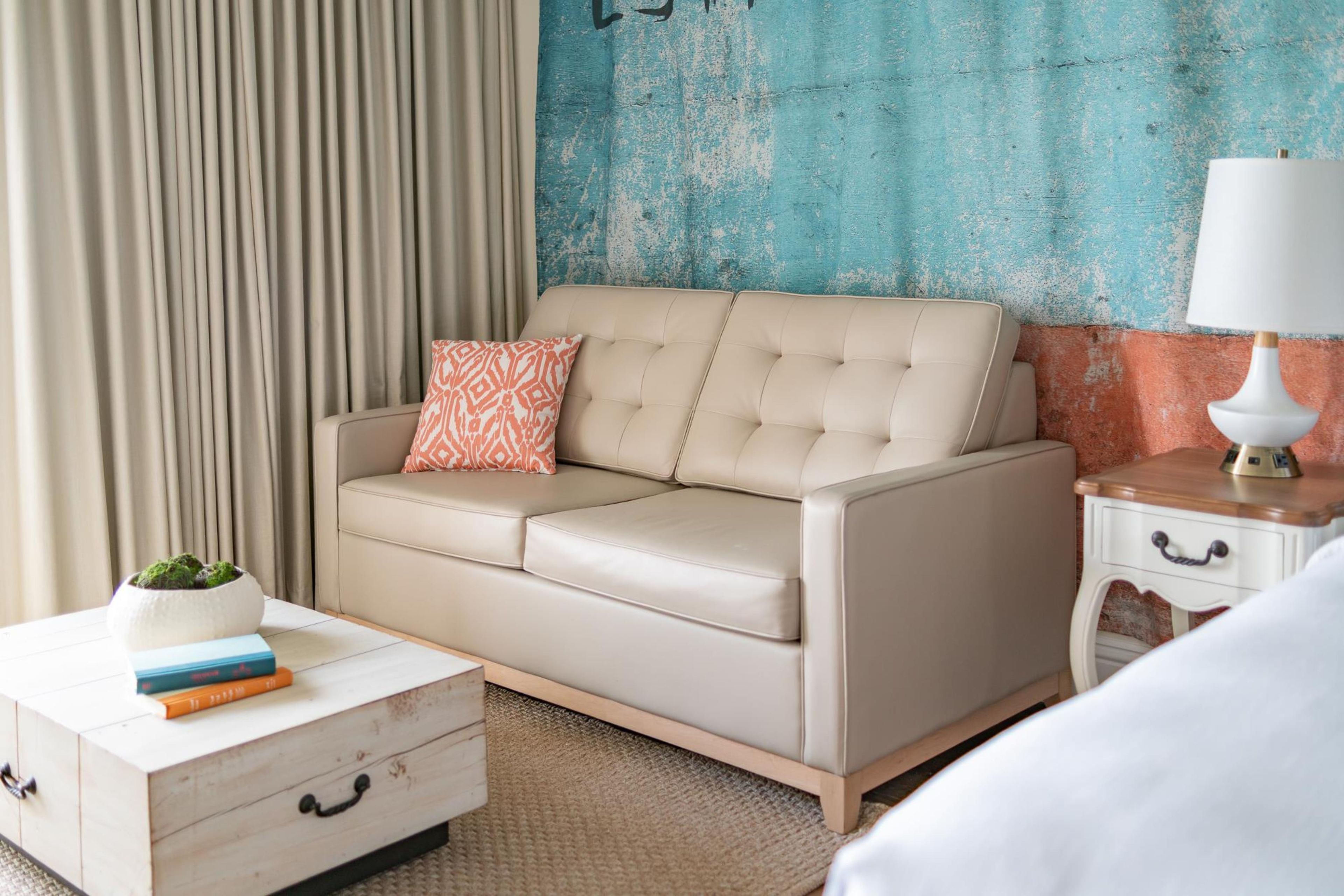 Habitación de hotel con un sofá color beige, una almohada estampada, una mesita de noche con una lámpara y una planta pequeña sobre un tocador blanco.