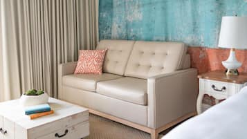 Habitación de hotel con un sofá color beige, una almohada estampada, una mesita de noche con una lámpara y una planta pequeña sobre un tocador blanco.