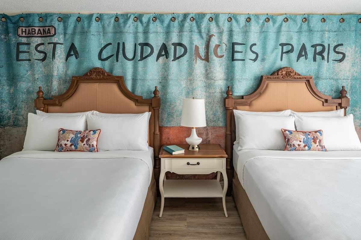 A hotel room with two beds, a nightstand with a lamp, and a wall with a sign reading "HABANA ESTA CIUDADES PARIS".