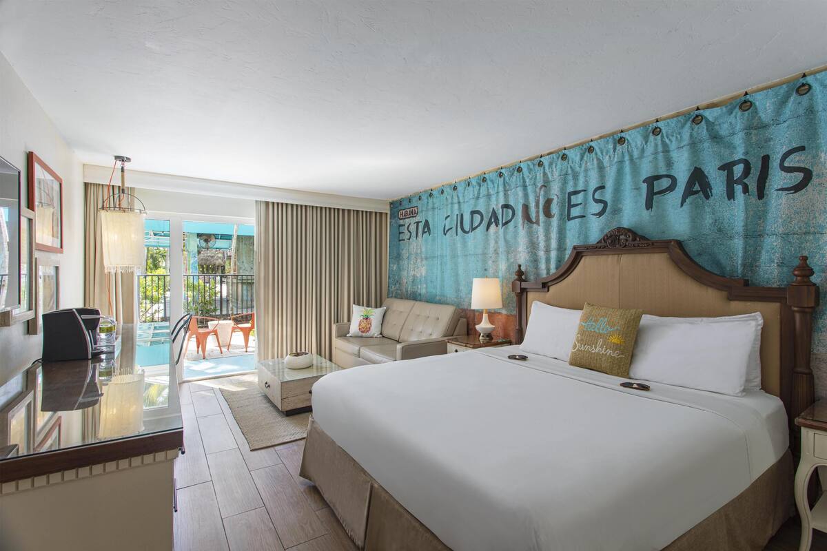 A hotel room with a large bed, a desk, a chair, a lamp, and a view of the outdoors.