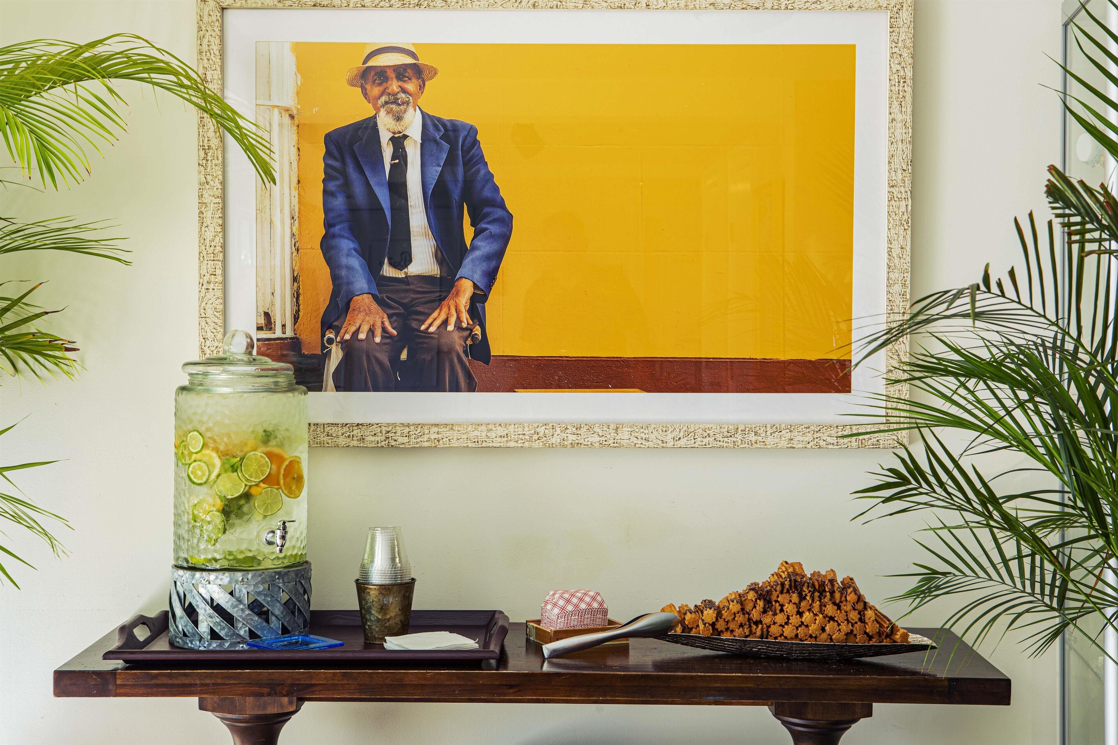 A framed portrait of a man in a blue suit, a glass jar with lime slices, a small metal container, and a tray with food items on a wooden table.