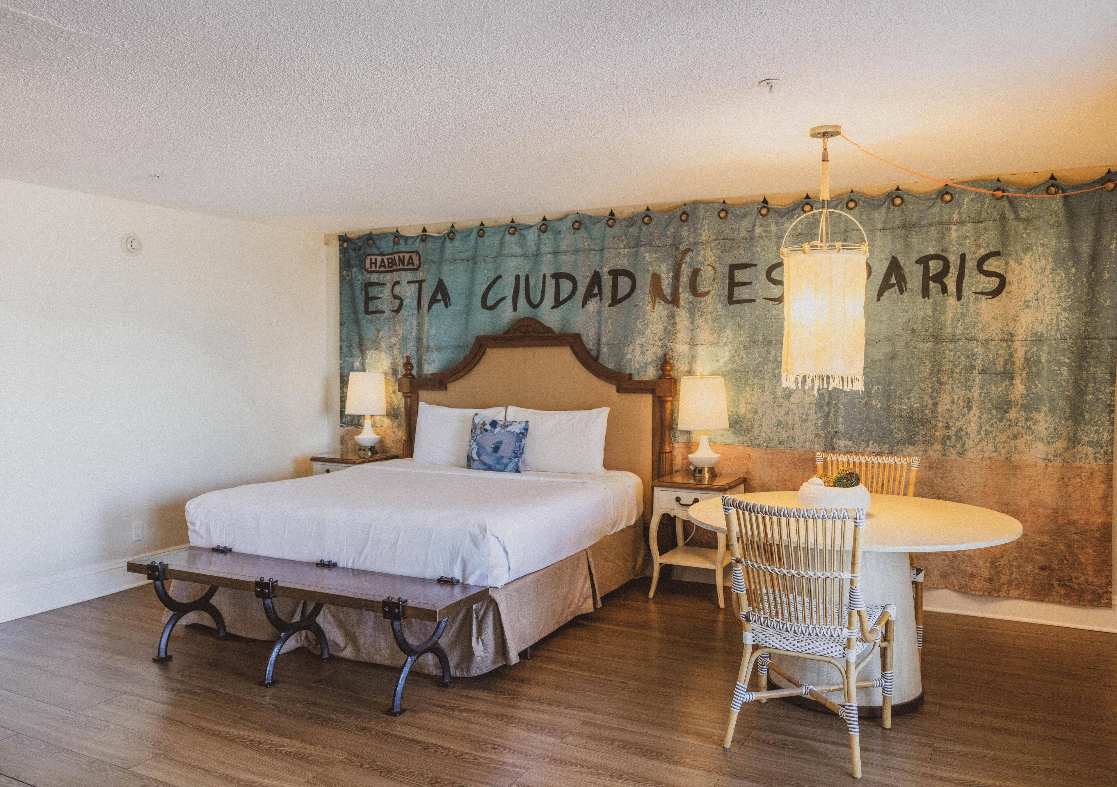 A hotel room with a bed, a table, and chairs. There is a curtain with text on the wall.
