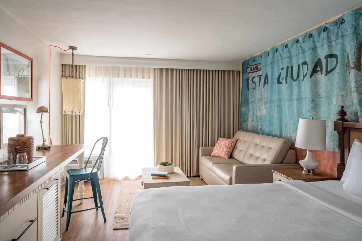 A hotel room with a bed, a desk, a chair, a sofa, and a large window with curtains.