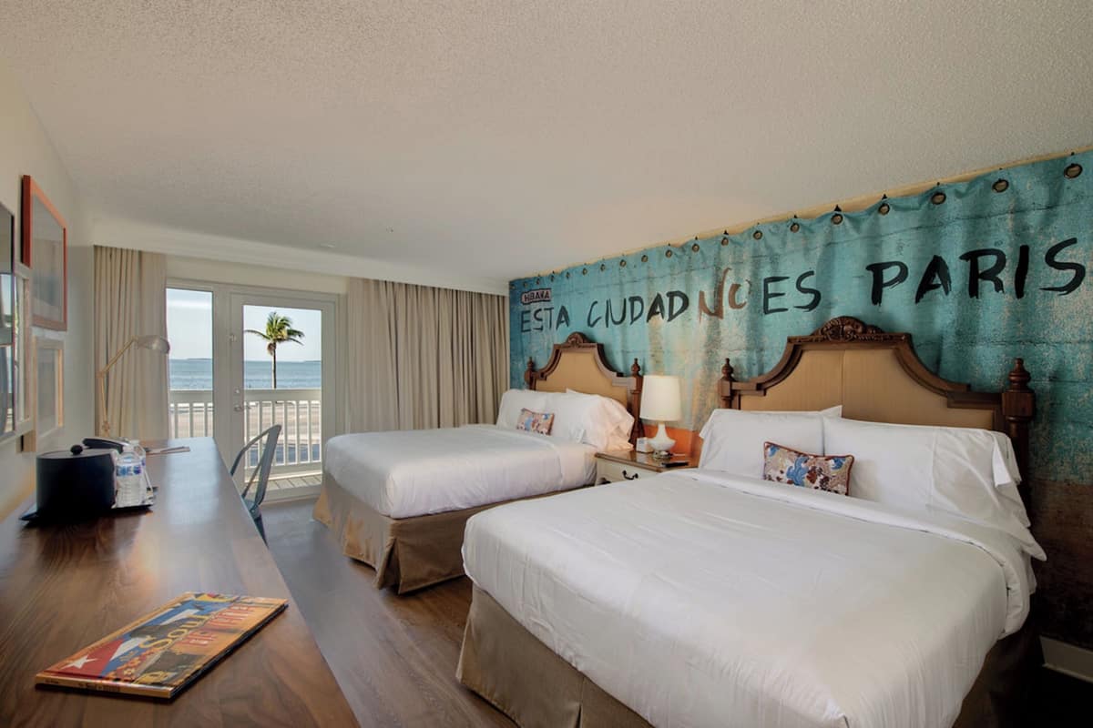 A hotel room with two beds, a view of the ocean, and a wall with 'ESTA CIUDAD NACES PARIS' text.