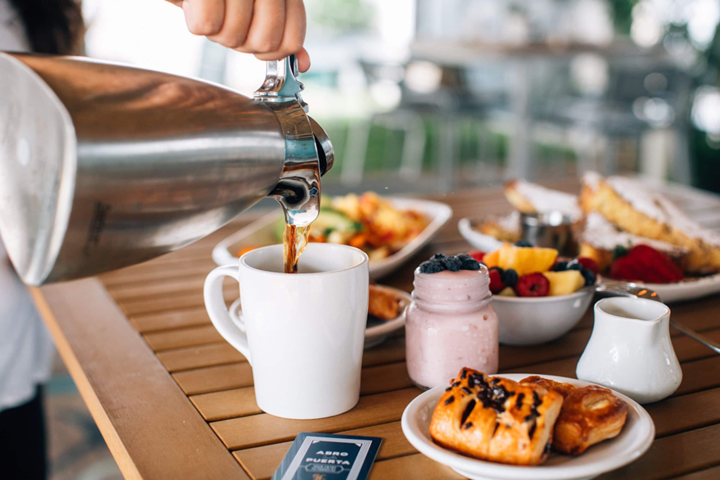 Un desayuno con una cafetera sirviendo café en una taza, un plato de pasteles, un bol de fruta y un tarro de mermelada sobre una mesa de madera.