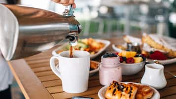 Un desayuno con una cafetera sirviendo café en una taza, un plato de pasteles, un bol de fruta y un tarro de mermelada sobre una mesa de madera.