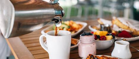 A breakfast setting with a coffee pot pouring into a mug, a plate of pastries, a bowl of fruit, and a jar of jam on a wooden table.