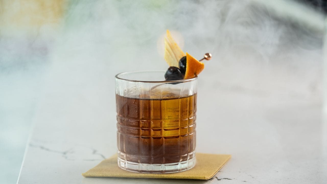 A glass of amber-colored beverage with a garnish, placed on a napkin on a marble surface.