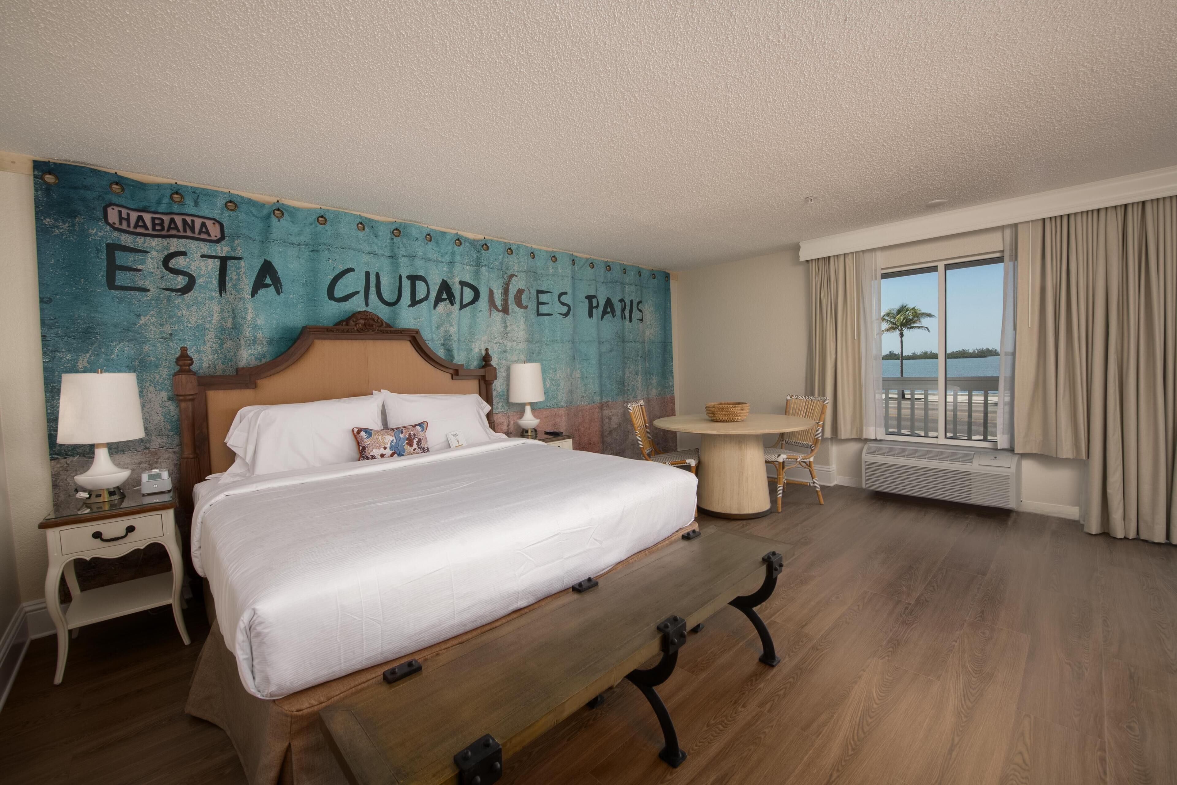 Habitación de hotel con una cama grande, dos mesitas de noche con lámparas, una mesa redonda y vistas al agua a través de amplias ventanas.
