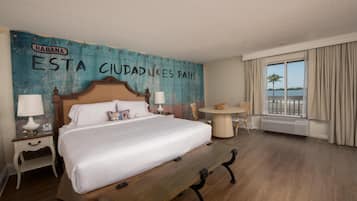 Habitación de hotel con una cama grande, dos mesitas de noche con lámparas, una mesa redonda y vistas al agua a través de amplias ventanas.