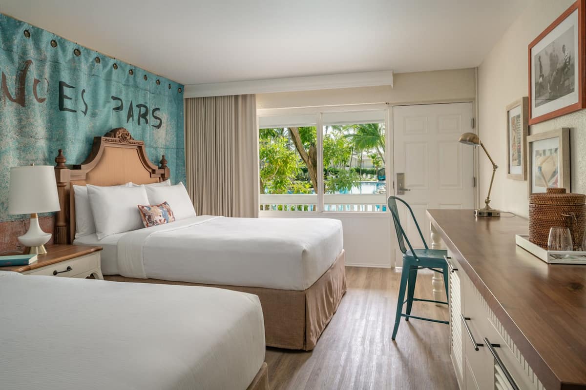 A hotel room with two beds, a desk, a chair, a window with a view of greenery, and a wall with 'NICE PARIS' written on it.