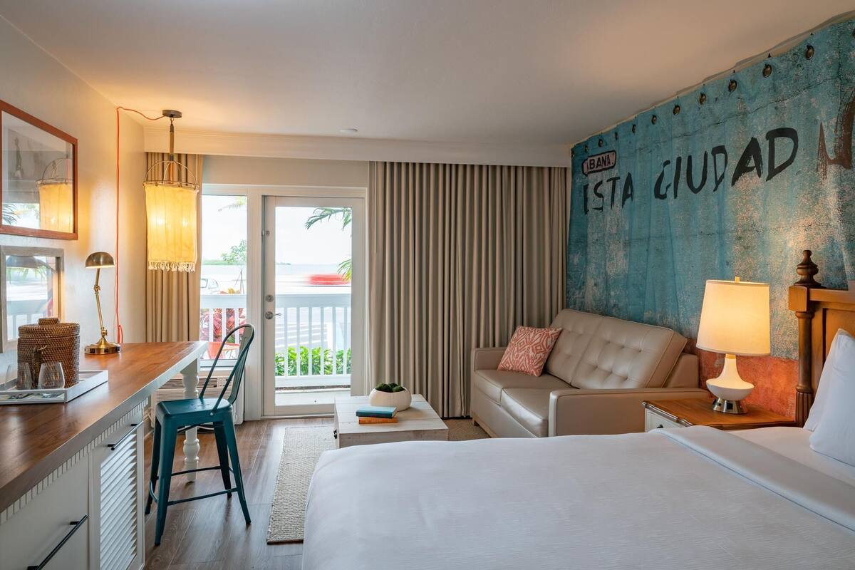 A hotel room with a bed, a sofa, a desk with a lamp, a chair, and a view of the outside.