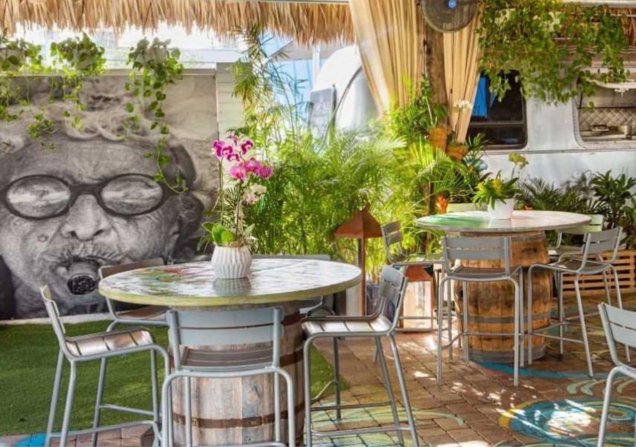 An outdoor seating area with round tables and metal chairs, surrounded by lush greenery and a mural.