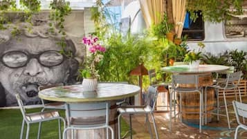 An outdoor seating area with round tables and metal chairs, surrounded by lush greenery and a mural.
