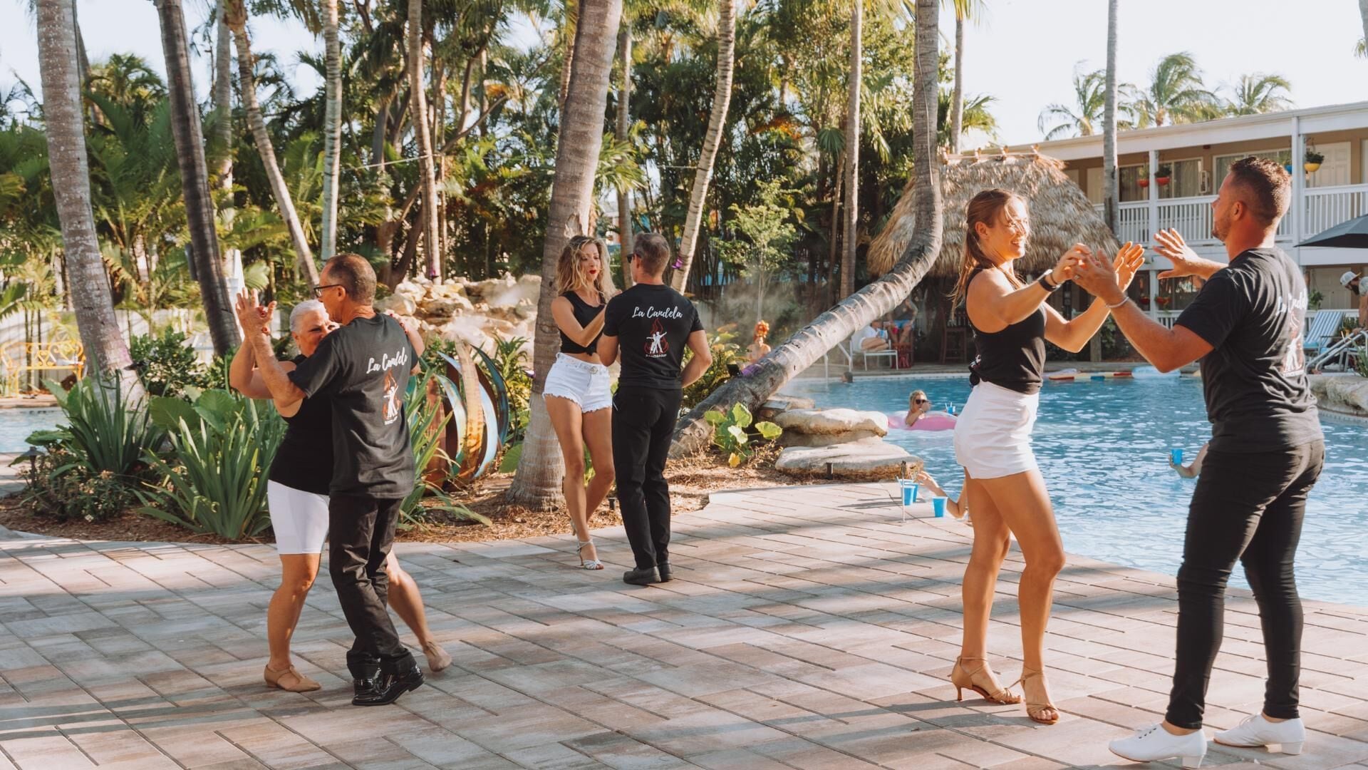 A group of people dancing by a pool with palm trees and a building in the background.