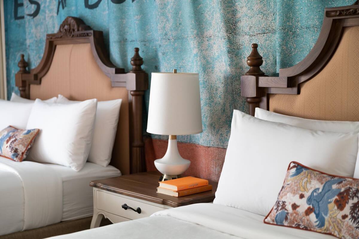 A hotel room with two beds, each with white linens and decorative pillows. There is a wooden nightstand with a lamp and books between the beds.