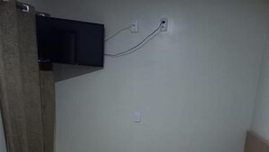 Un llit individual amb estovatge blanc i una manta daurada, una tauleta de nit de fusta, un televisor fixat a la paret i un assecador tovalloles amb una tovallola.