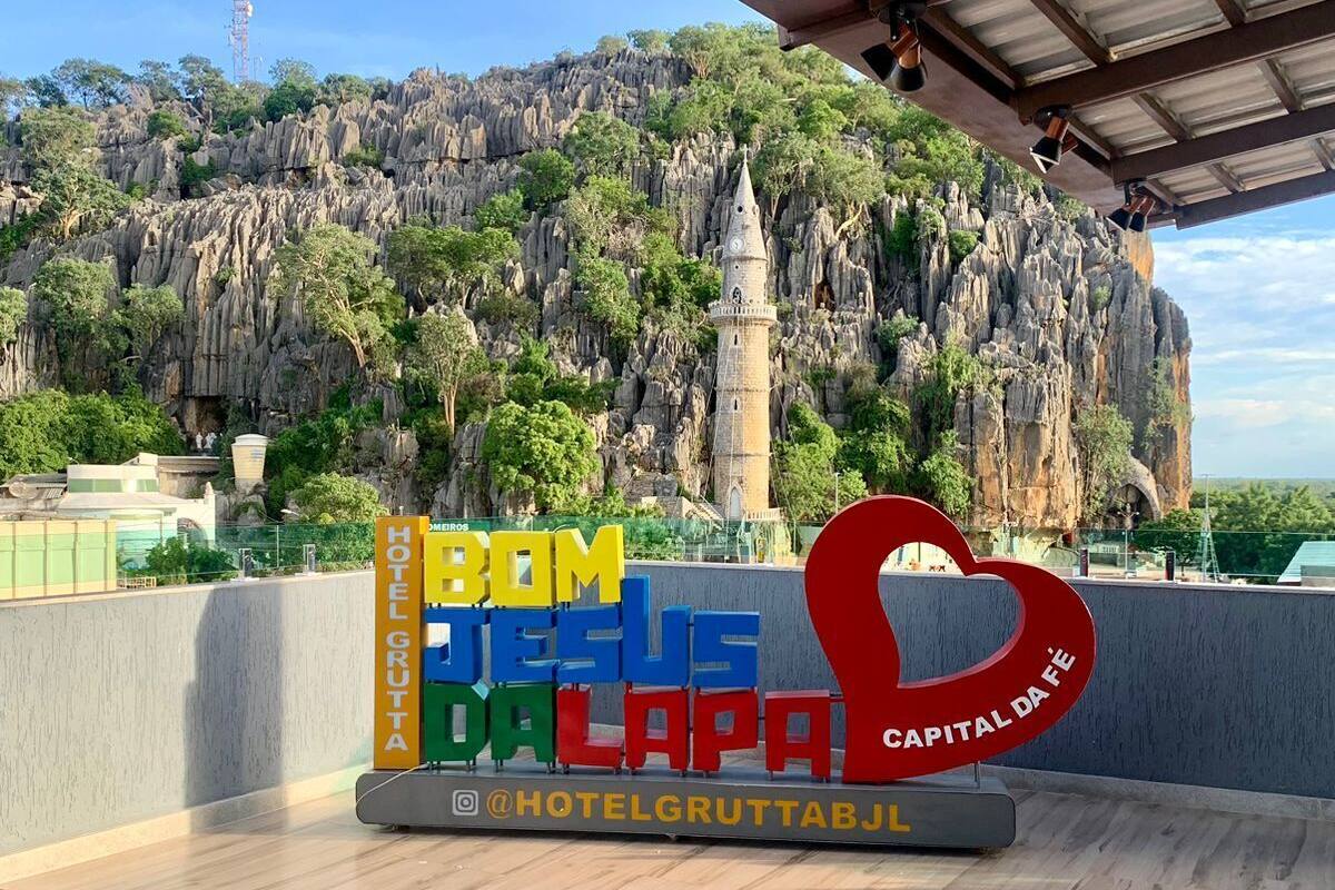 A hotel sign with 'HOTEL GRUTTA' and 'BOM JESUS DA LAPA' in colorful letters, a heart-shaped logo, and a view of a rocky landscape with trees.