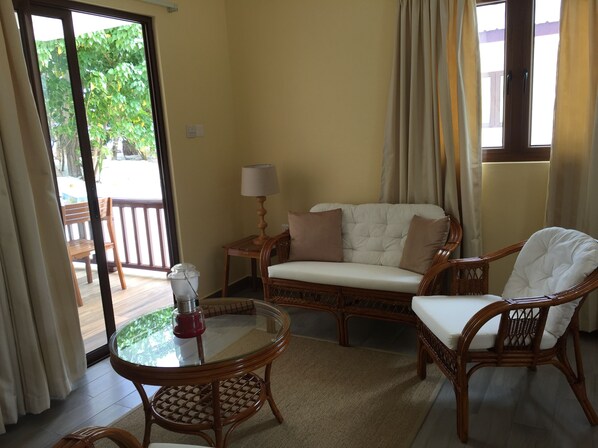 A living room with a sofa, two chairs, a glass-top table, a lamp, and a view through a glass door to an outdoor area with a bench.