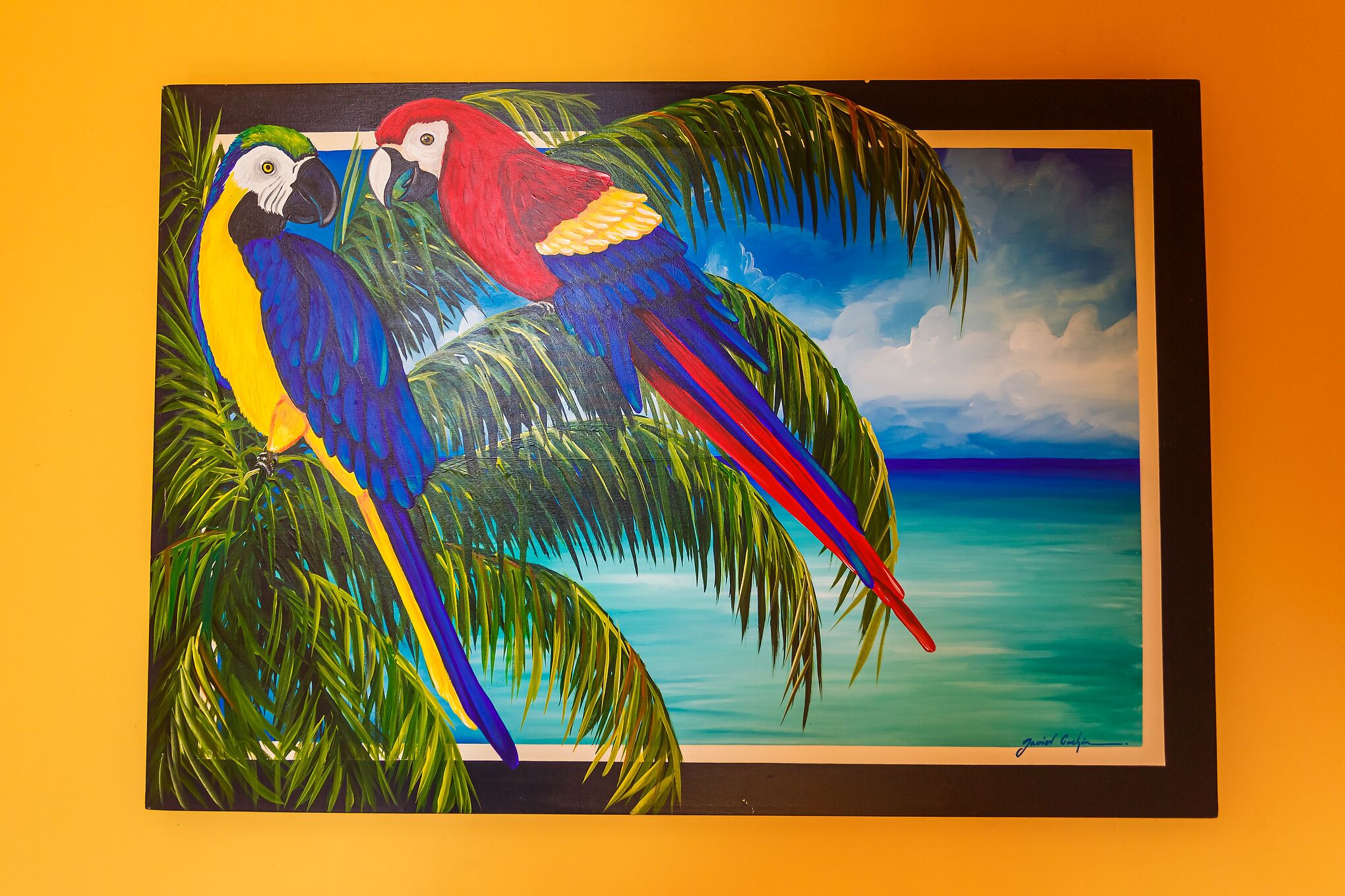 Un tableau encadré représentant deux perroquets aux couleurs vives, perchés sur des feuilles de palmier, avec un paysage tropical en arrière-plan.