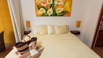 A neatly made bed with white and beige bedding, a painting of flowers on the wall, and a wooden basket with towels on the side.