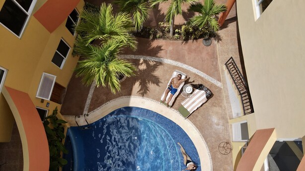An indoor swimming pool with a tiled deck, surrounded by palm trees and a building with a curved facade. - Kaam Accommodations (Puerto Morelos)