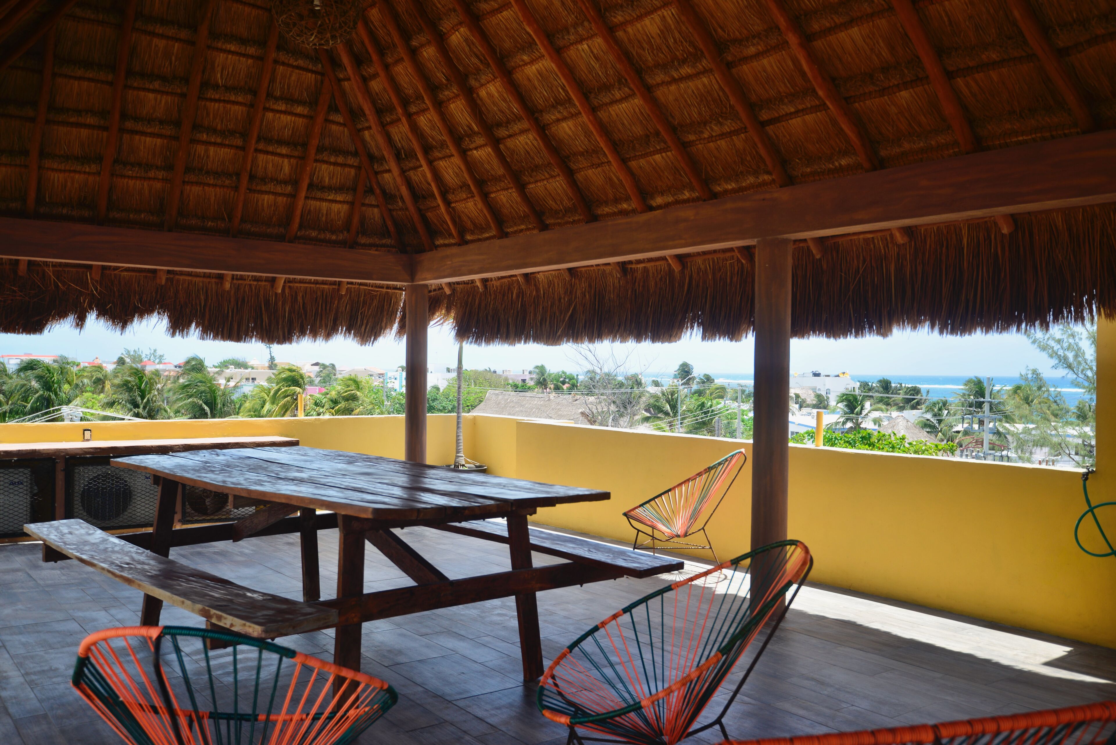 A covered patio with a wooden table and chairs, a thatched roof, and a view of the ocean and palm trees.