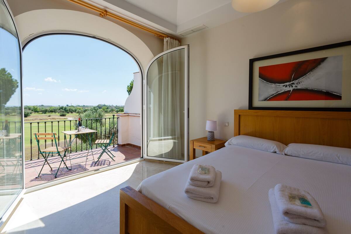 Habitación de hotel con una cama grande, un cuadro, una mesita de noche, una lámpara y vistas a un jardín a través de un ventanal.