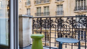 A balcony with a green stool and a small table, overlooking a building with ornate balconies and windows. - CMG Tour Eiffel (Paris)
