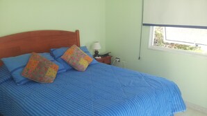 A bed with blue bedding and colorful pillows, a wooden headboard, a bedside table with a lamp, and a window with a view of greenery. - Casa en Bahia Inglesa (Caldera)