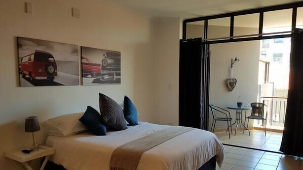 Ein Hotelzimmer mit einem Bett, einem kleinen Tisch mit Lampe, einem Balkon mit Tisch und Stühlen sowie zwei an der Wand befestigten Bildern von Autos.