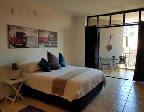 Quarto de hotel com cama, uma pequena mesa com abajur, varanda com mesa e cadeiras, e duas imagens de carros penduradas na parede.