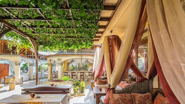 Een terras met tafels en banken, overdekt door een pergola met klimplanten.