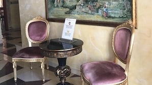 Een ontvangstruimte met twee sierlijke stoelen, een tafel en een groot klassiek schilderij van oude ruïnes.